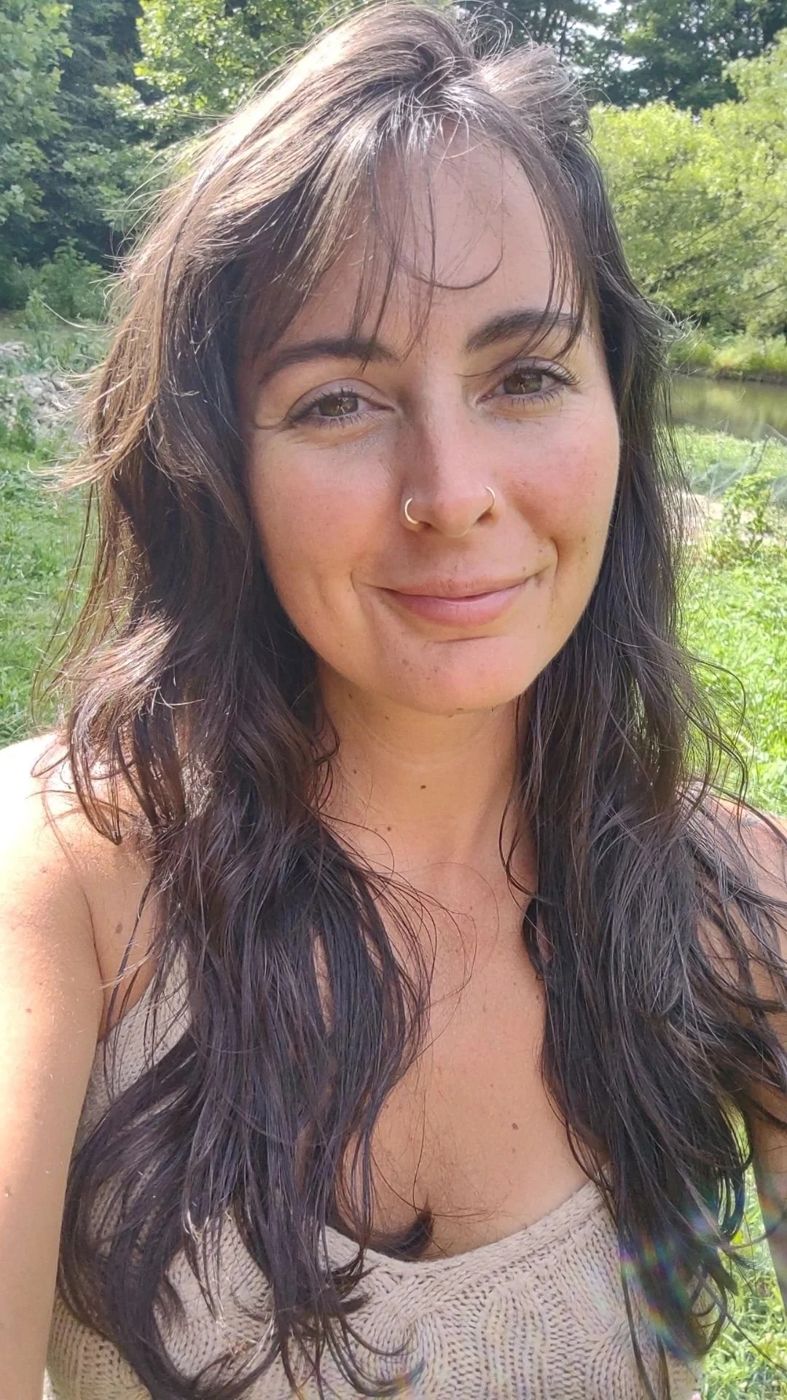 Alisha smiling outdoors with green trees and sunlight in the background